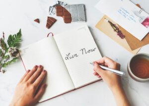 Ein aufgeschlagenes Notizbuch auf dessen Seite "Live now" geschrieben steht. Frauenhände, die einen Stift halten. Um das Notizbuch herum liegen Blumen, eine angebrochene Schokoladentafel, einige Briefe und eine Tasse Tee. Journaling und Achtsamkeit gegen Stress.