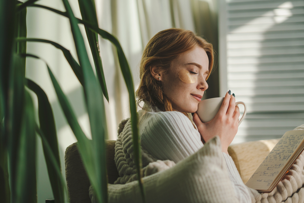 Pretty dreamy young woman sitting on couch covered with a blanket, drinking coffee indoors at home room. Cosy scene, cozy home concep. Stress reduction. Mindfulness, Selfcare.