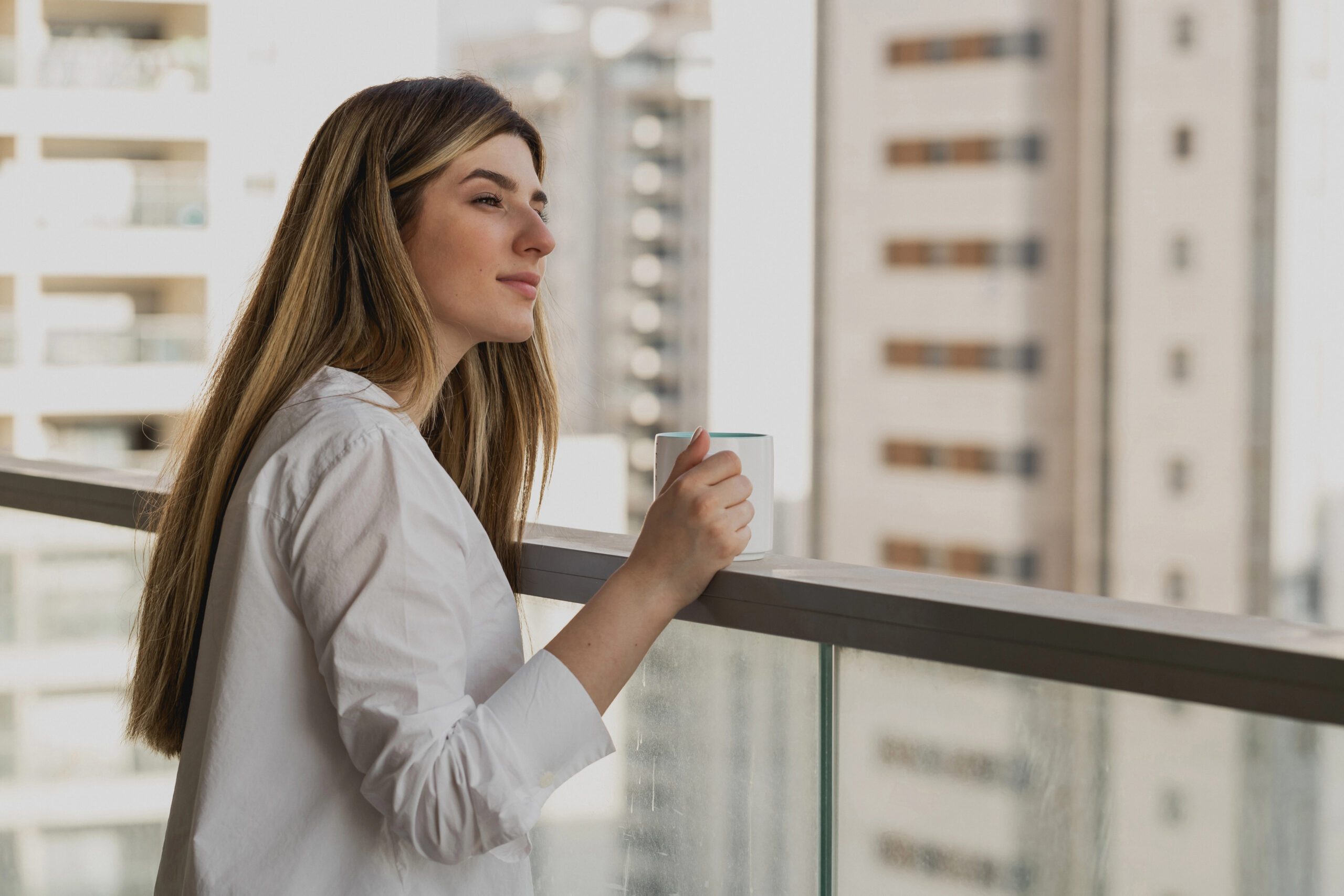 Frau mit Kaffetasse steht auf dem Balkon, macht Pause und schaut in die Ferne. 