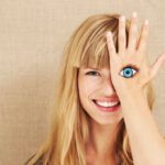 Eine lachende, blonde Frau mit strahlenden Augen, die mit einer Hand ihr linkes Auge verdeckt. Auf dem Handrücken ist ein Auge aufgemalt.