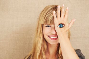 Eine lachende, blonde Frau mit strahlenden Augen, die mit einer Hand ihr linkes Auge verdeckt. Auf dem Handrücken ist ein Auge aufgemalt.