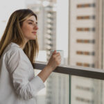 Frau mit Kaffetasse steht auf dem Balkon und schaut in die Ferne.