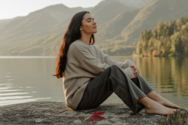 Eine dunkelhaarige Frau sitzt mit geschlossenen Augen am Ufer eines Sees. Im Hintergrund Wald. Herbststimmung.